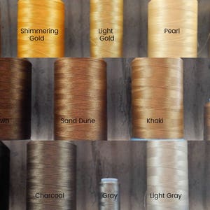 以下が含まれることがあります： Old Gold、Brown、Whiteなどの色でラベル付けされた、ニュートラルな色調の糸巻きのコレクション。画像には、輝くゴールド、カーキ、ライトグレーなど、さまざまな色合いが表示されています。「Fine Thread Art Neutrals」というテキストが表示されています。