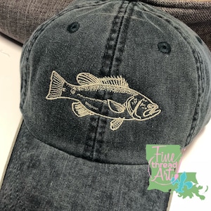 May include: A dark blue baseball cap with a beige embroidered bass fish design. The cap has a curved brim and a white stitching detail around the brim. The text "Fine thread Art" is embroidered on the side of the cap.