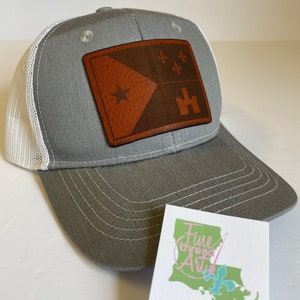 May include: Gray and white trucker hat with a brown leather patch featuring the New Orleans flag. The patch has a white star, three fleur-de-lis, and a brown castle. The hat has a white mesh back and a gray front. The hat is sitting on a white surface next to a business card that says "Fine Thread Art Embroidery".