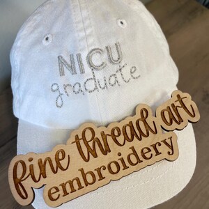 May include: White baseball cap with the text "NICU graduate" embroidered in silver thread. A wooden sign with the text "fine thread art embroidery" is attached to the brim of the cap.