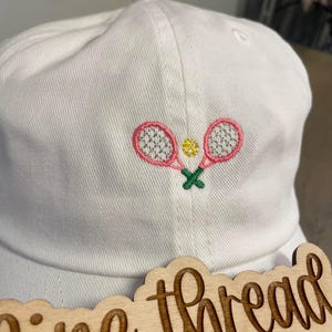 May include: White baseball cap with pink and green embroidered tennis rackets and a yellow tennis ball. The cap is sitting on a wooden sign that says "Fine thread embroidery".
