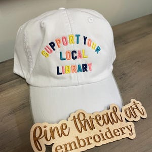 May include: White baseball cap with the text "Support Your Local Library" embroidered in rainbow colors. A wooden tag with the text "fine thread art embroidery" is in the foreground.