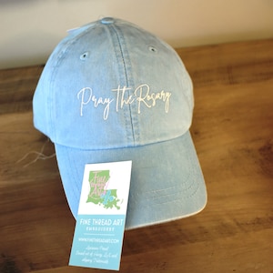 May include: A light blue baseball cap with white embroidery that reads "Pray the Rosary". The cap is on a wooden surface with a business card that reads "Fine Thread Art Embroidery" and a website address.