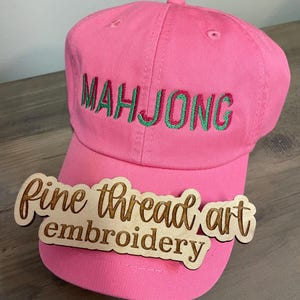 May include: A bright pink baseball cap with the word "MAHJONG" embroidered in green thread. A wooden sign that says "Fine thread art embroidery" is in front of the cap.