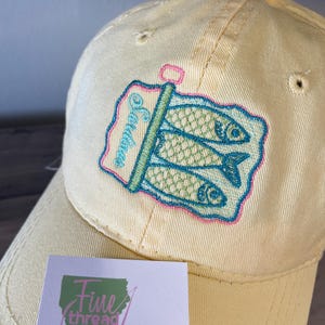 以下が含まれることがあります： 刺繍されたイワシ缶のデザインが施された、薄い黄色の野球帽。デザインは、3匹の緑色の魚、ターコイズ色の缶切り、ピンク色の「Sardines」の文字が特徴です。「Fine thread Art」というテキストの小さなカードが前景にあります。