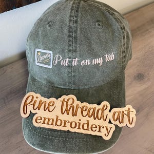 May include: An olive green baseball cap featuring an embroidered white square logo with the word Library and a small gold heart, followed by the embroidered white text Put it on my tab. A wooden sign with the text fine thread art embroidery is placed over the brim.