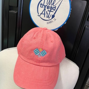 May include: A pink baseball cap with an embroidered design of two blue and white paddleball rackets and a yellow ball. The cap has a curved brim and a white stitching detail around the brim.