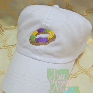 May include: White baseball cap with an embroidered Mardi Gras king cake design. The king cake embroidery features green, yellow, and purple icing on a brown cake base. The cap has a curved brim and a button on top.