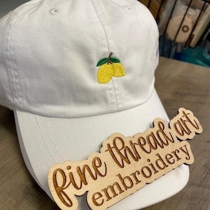 May include: A white baseball cap with an embroidered lemon design. The cap features a curved brim and a small, embroidered image of two yellow lemons with green leaves. A wooden sign with the text "Fine thread art embroidery" rests on the cap.