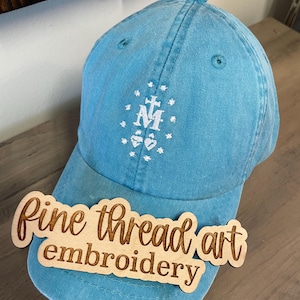 May include: A light blue denim baseball cap with white embroidery of the Miraculous Medal. The cap has a wooden tag attached to the brim that says "fine thread art embroidery".