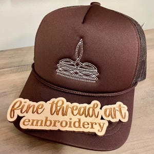 May include: A brown trucker hat with a white embroidered design and a mesh back. The hat has a wooden sign that reads "Fine thread art embroidery". The hat is sitting on a wooden surface.