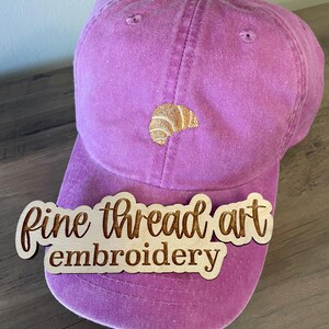 May include: A dusty rose baseball cap with a curved brim. The cap features an embroidered croissant design in shades of tan and brown. A wooden sign that reads "Fine thread art embroidery" is placed on the cap.