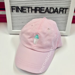 May include: A pink baseball cap with an embroidered design of a blue and green snow cone in a white cup. The cap has the text "FINETHREADART" embroidered on the side.
