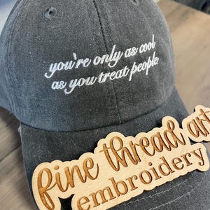 May include: A charcoal gray baseball cap featuring white embroidered text that reads, you're only as cool as you treat people. A light-colored wooden cutout with the engraved text Fine thread art embroidery is positioned across the brim.