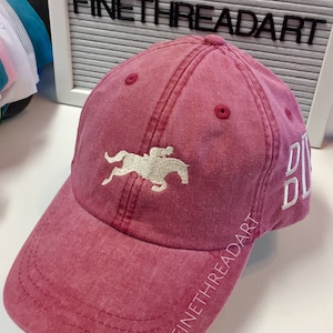 May include: A red baseball cap with a white embroidered image of a horse and rider. The text "FINETHREADART" is embroidered on the side of the cap.