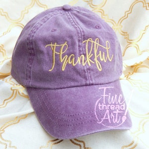 May include: Purple baseball cap with the word "Thankful" embroidered in gold thread. The cap has a curved brim and a logo on the side that says "Fine thread Art".