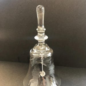 May include: Clear glass bell with a long handle and a flared base. The bell features etched grape and leaf designs. A small, clear crystal hangs from a chain inside the bell. The bell is likely used for decorative purposes.
