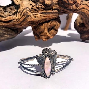 May include: A silver cuff bracelet with a central pink oval stone. The bracelet features decorative leaf and floral elements. The design includes twisted wire and a detailed setting. The background includes a piece of driftwood.