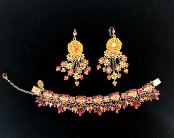 Può includere: Un set di gioielli con bracciale e orecchini abbinati. Gli orecchini e il bracciale sono adornati con motivi floreali gialli, arancioni e rossi e dettagli pendenti. Il set è presentato su sfondo nero.