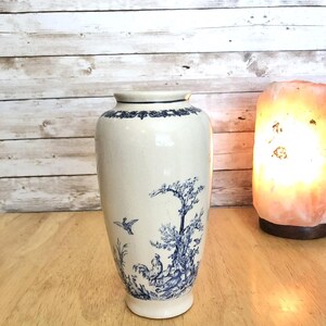 May include: A white ceramic vase with a blue floral and bird design. The vase has a cylindrical shape with a slightly flared base and a detailed blue border around the top. The design features a bird in flight and a tree.