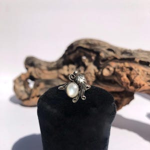 May include: A silver ring with a pearlescent oval stone, accented by leaf and swirl designs. The ring is displayed on a black velvet surface against a backdrop of driftwood and a white background. The ring's design is intricate and detailed.