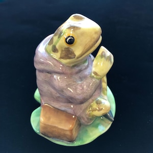 May include: A ceramic frog figurine, featuring a light yellow head with brown spots, wearing a purple robe. The frog is holding a yellow object and sitting on a green lily pad. A brown rectangular object is at its side.