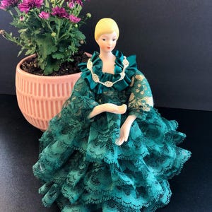 May include: A porcelain doll with blonde hair and a teal lace dress. The doll has a white collar and is posed with arms outstretched. A pink potted plant with purple flowers is in the background.