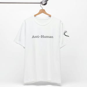 Könnte beinhalten: Weißes T-Shirt mit dem schwarzen Aufdruck "Anti-Human". Das Shirt hängt an einem Holzbügel. Auf dem rechten Ärmel ist eine kleine Grafik aufgedruckt. Das Shirt ist aus weichem Material.