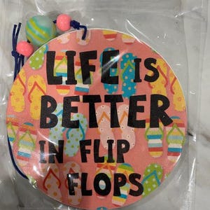 May include: A round, pink air freshener with the text "LIFE IS BETTER IN FLIP FLOPS" in black. The design includes colorful flip-flop illustrations. The air freshener is strung with a blue cord and decorative beads.