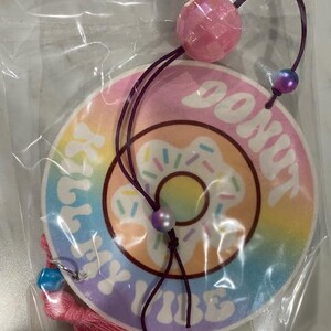 May include: A round air freshener with a donut design, featuring the text "DONUT KILL MY VIBE". The air freshener has a pink, purple, and yellow color scheme, with a pink tassel and a purple string with beads.