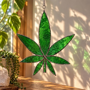 Op de afbeelding: Een glas-in-lood suncatcher in de vorm van een cannabisblad met zeven punten. Het object is gemaakt van getextureerde, levendig groene glaspanelen die bijeengehouden worden door donkere metalen soldeerlijnen, met een metalen lus om op te hangen.