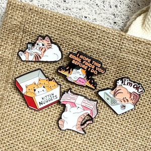 May include: A selection of cat-themed enamel pins displayed on a hessian tote bag. The pins depict various cat illustrations, including a box of "Kitten Nuggets" and a cat with the text "I'm ok?". Other pins have text that reads "I hope this email finds you before I do."