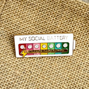 May include: A rectangular enamel pin with the text "MY SOCIAL BATTERY" at the top. The pin features a battery level indicator with various emoji faces, ranging from sad to happy, and a lightning bolt.