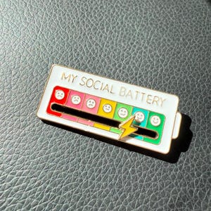 May include: A white enamel pin with the text "MY SOCIAL BATTERY" and a colorful battery level indicator. The indicator features a range of emoji faces from sad to happy, with a yellow lightning bolt. The pin is on a textured surface.