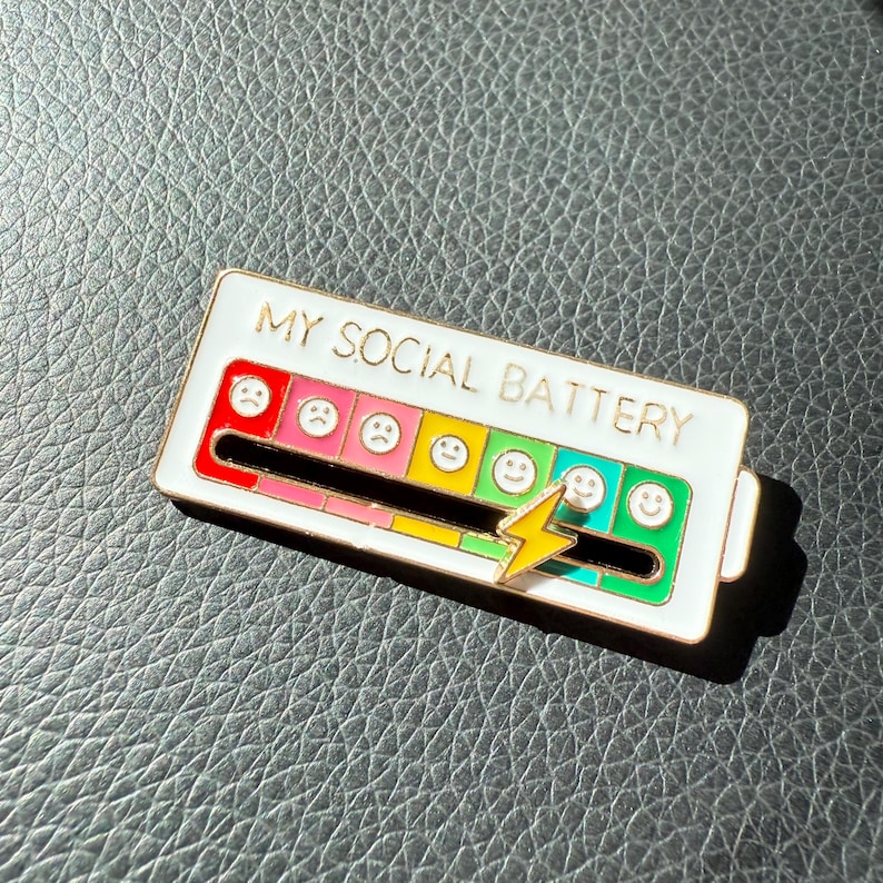May include: A white enamel pin with the text "MY SOCIAL BATTERY" and a colorful battery level indicator. The indicator features a range of emoji faces from sad to happy, with a yellow lightning bolt. The pin is on a textured surface.