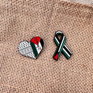 Puede incluir: Dos pines esmaltados con la bandera palestina. Un pin tiene forma de corazón con un patrón en blanco y negro y los colores de la bandera. El otro pin es una cinta con los colores de la bandera. Ambos pines están sobre una superficie texturizada y beige.