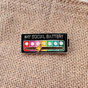 Puede incluir: Un pin de esmalte negro con el texto "MY SOCIAL BATTERY" encima de un indicador de nivel de batería. El indicador muestra una gama de emociones, de triste a feliz, con un rayo amarillo. El pin está sobre una superficie texturizada y beige.