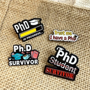 Könnte beinhalten: Vier schwarze Emaille-Anstecknadeln mit weißem Text und Grafiken. Eine Anstecknadel lautet "PhD IN PROGRESS..." mit einer Grafik einer Graduiertenkappe. Eine andere sagt "Trust me I have a PhD". Die anderen beiden Anstecknadeln lauten "Ph.D SURVIVOR" und "PhD Student SURVIVOR".