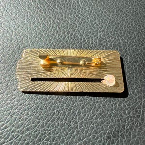 May include: A gold-colored rectangular pin with a sunburst design. The pin has a safety clasp on the back and a clear, round plastic piece. The pin is on a black leather-like surface.