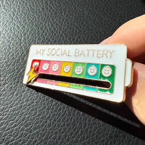 May include: A white enamel pin with the text "MY SOCIAL BATTERY". The pin features a battery level indicator with a lightning bolt and a series of emoji faces ranging from sad to happy, indicating social energy levels.