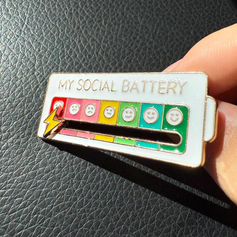 May include: A white enamel pin with the text "MY SOCIAL BATTERY". The pin features a battery level indicator with a lightning bolt and a series of emoji faces ranging from sad to happy, indicating social energy levels.