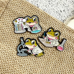 Peut inclure: Trois épingles en émail représentant des grenouilles de dessin animé en blouses de laboratoire. Une grenouille utilise un microscope, une autre écrit et la troisième travaille avec des tubes à essai. Les épingles sont blanches, vertes, roses et noires.