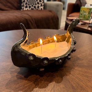 May include: A handcrafted, dark green ceramic candle holder shaped like a Viking ship. The interior is filled with sand and two lit wicks. The candle holder has decorative details and is placed on a wooden surface.