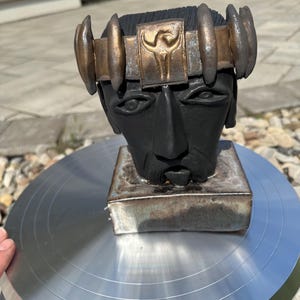 Könnte beinhalten: Schwarze Keramikbüste eines Mannes mit bronzefarbener Krone. Die Büste ist auf einem quadratischen Sockel mit metallischem Finish montiert. Die Krone hat ein zentrales Emblem und ist mit abgerundeten Akzenten verziert.