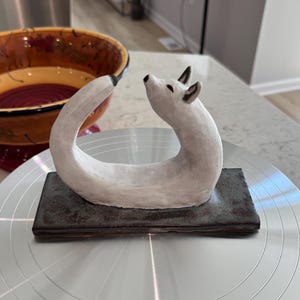 Puede incluir: Escultura de zorro de cerámica blanca con cuerpo curvado y cabeza levantada, sobre una base rectangular gris oscuro. Las orejas y la punta de la cola del zorro son negras. La escultura se exhibe sobre una superficie plateada.