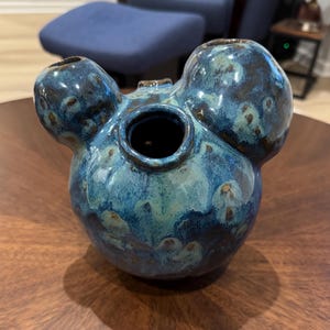 May include: A ceramic vase with a unique shape and a glossy, blue glaze. The vase has a central opening and two bulbous protrusions on either side, with a third on top. The glaze features shades of teal, blue, and brown.