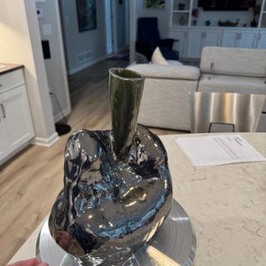 May include: A unique, handcrafted ceramic vase with a dark, metallic finish. The vase has an organic, sculptural form with a dark green, cylindrical neck. The vase is displayed on a silver rotating stand, showcasing its reflective surface.