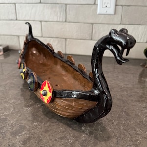 May include: A ceramic Viking ship planter with a black serpent head and shields. The ship is brown with black trim and features colorful shields along the side. The planter is designed to hold plants or other decorative items.