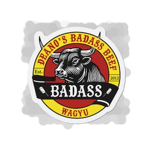 May include: A sticker with a black and white bull illustration, encircled by yellow and red text reading "Deano's Badass Beef". The word "Badass" is on a black banner, with "Wagyu" below. The design includes the year "2012".