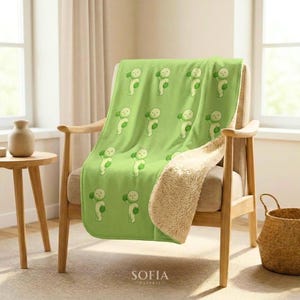 May include: A light green throw blanket featuring a repeating pattern of small, cartoonish figures holding green spheres. The blanket has a soft, textured cream-colored sherpa lining visible on the folded edge, draped over a wooden armchair.